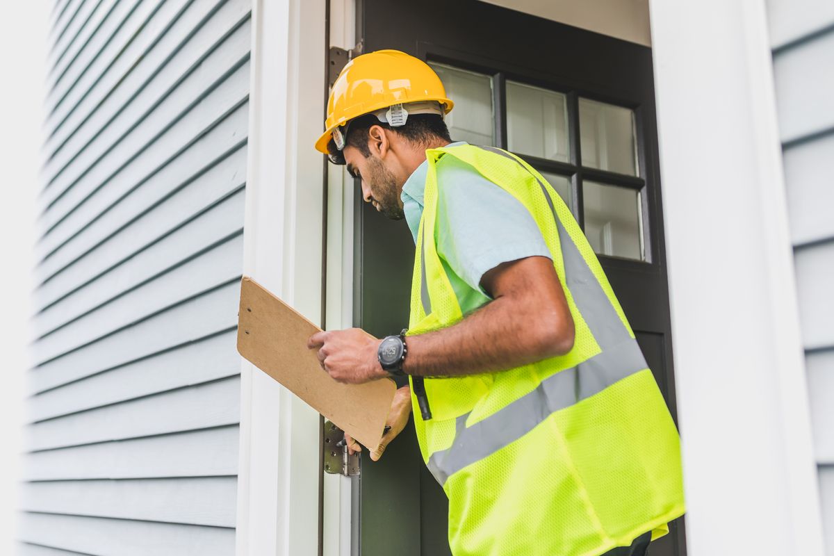 Inspector in a yellow hard hat and safety vest examining a building exterior doorway while holding a clipboard
