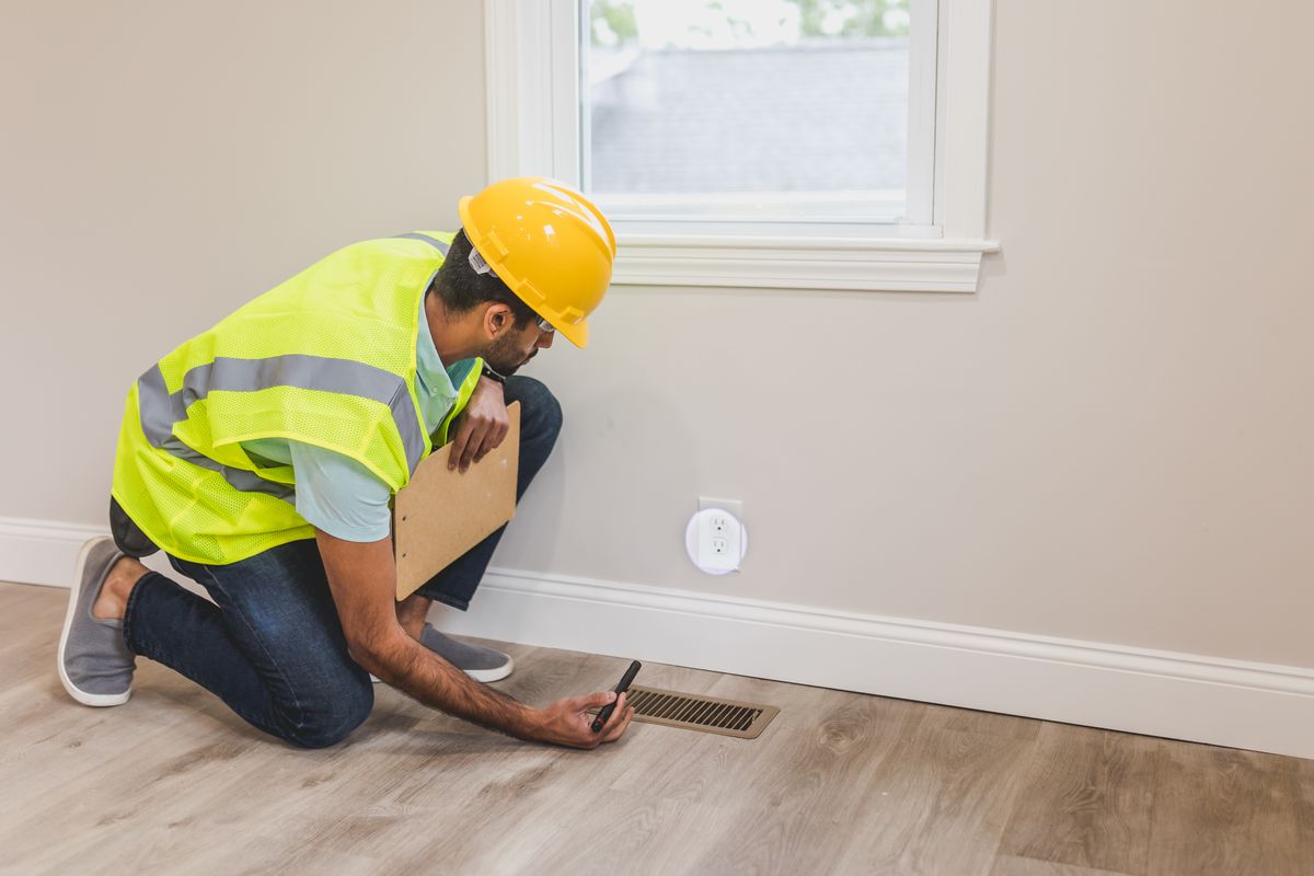 Inspector in a yellow hard hat kneeling on a wood floor, shining a small flashlight at a floor vent near the baseboard