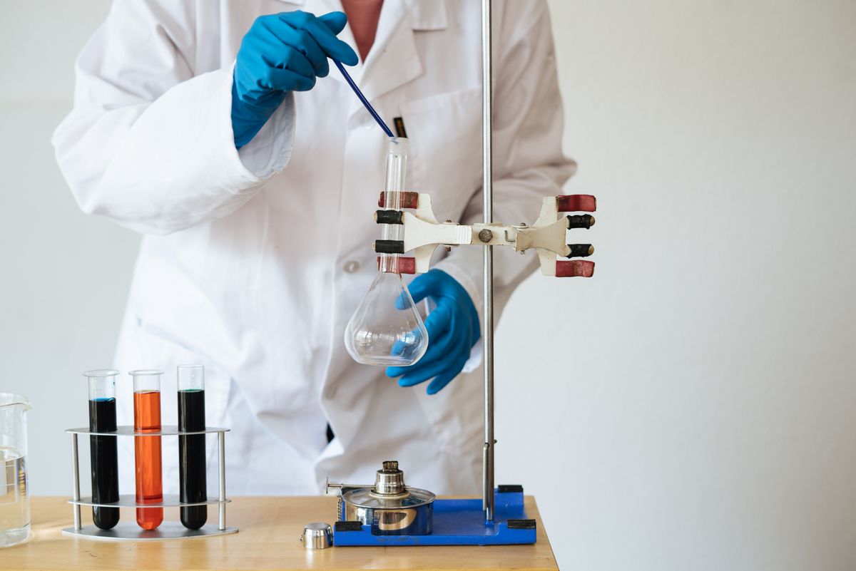 Laboratory technician in a white coat and blue gloves pipetting into a flask beside a rack of test tubes